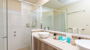 Ein modernes Badezimmer mit Duschkabine aus Glas, einem großen Spiegel und einem Waschtisch mit weißem Waschbecken und Pflegeprodukten.