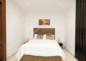 A bedroom with a large bed, two bedside tables, a painting above the bed, and a wooden floor. - The Lennox Suite by HDG (Accra)