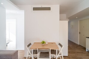 Ein moderner Essbereich mit einem Holztisch und weißen Stühlen, eine kleine Pflanze auf dem Tisch und eine weiße Wand mit Lüftungsgitter darüber.