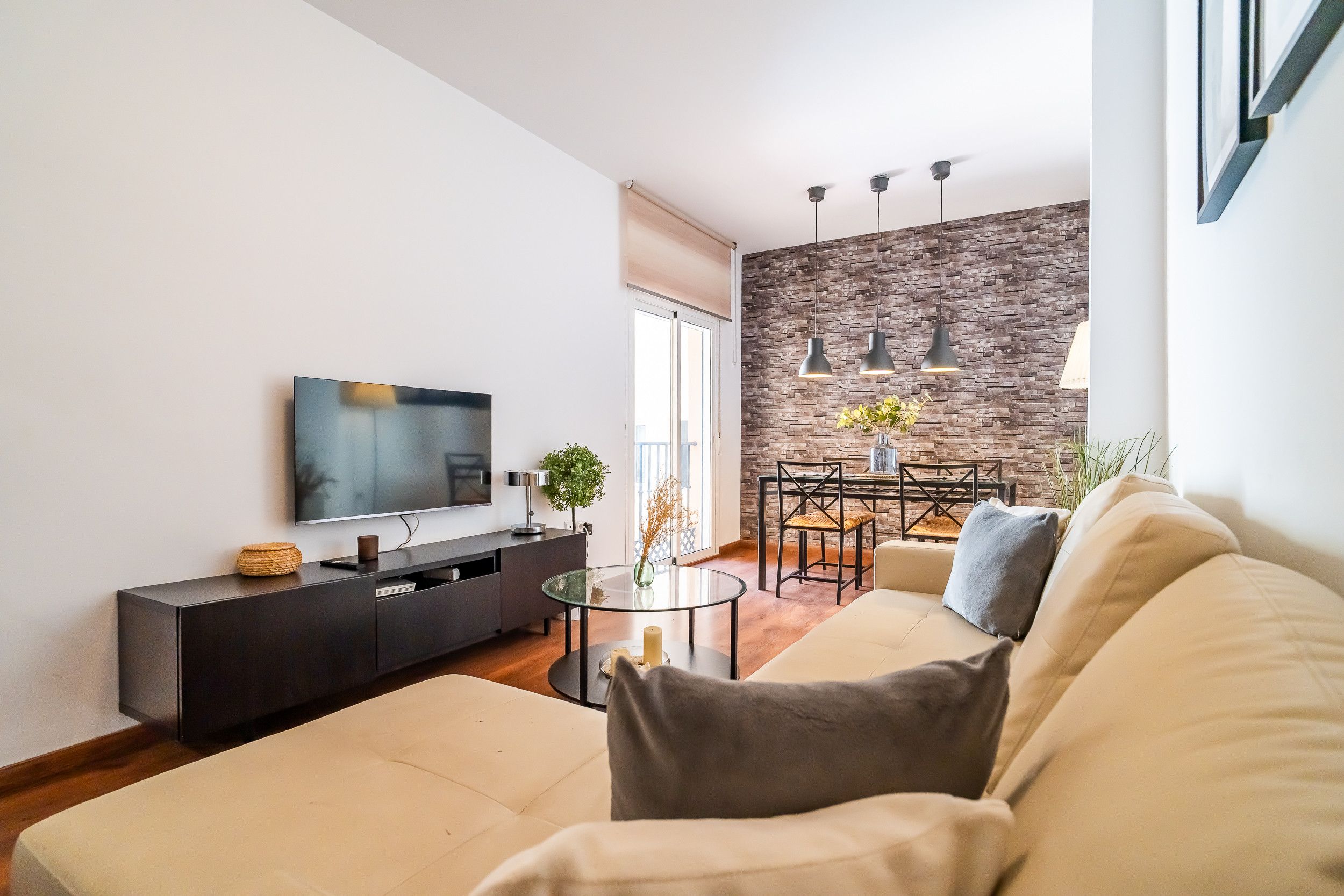 A modern living room with a flat-screen TV, a sofa, a dining area with a table and chairs, and a stone wall.