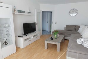 A living room with a television on a white entertainment unit, a grey sofa, a coffee table with a vase of flowers, and a clock on the wall. - Holiday home in Hage with swimming pool (Hage)