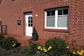 A red brick building with a white door and window shutters, number 3a, and a small garden with yellow flowers. - Holiday home in Hage with swimming pool (Hage)