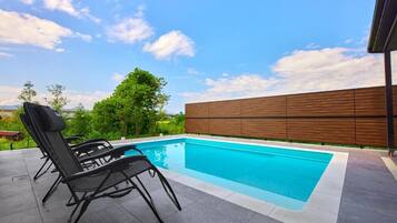 Une piscine entourée de chaises longues, délimitée par une clôture en bois et bordée de verdure.