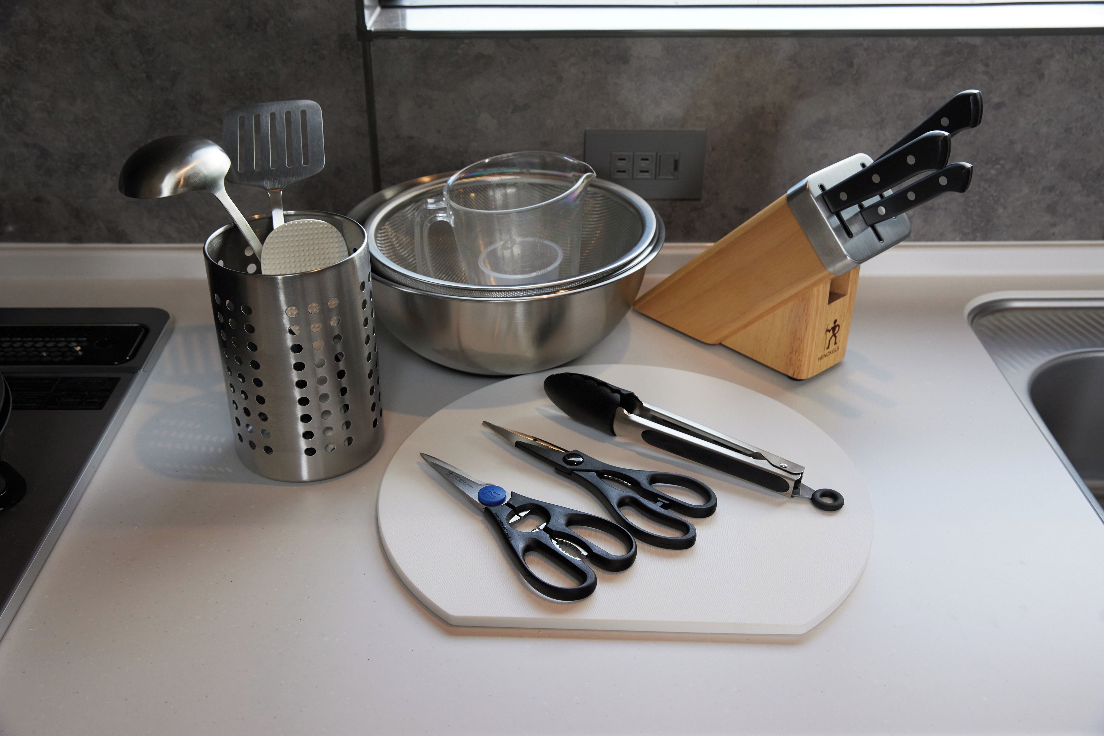 A kitchen counter with a colander, a strainer, a pair of scissors, and a knife block.