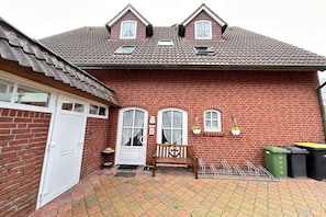 A red brick house with a white door, two windows with curtains, a bench, and a small garden area. - Holiday home Johren Apt. 2 (Norderney)