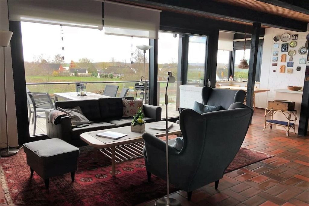 Una sala de estar moderna con un sofá, un sillón y un reposapiés, una mesa de comedor y un ventanal con vistas al exterior.