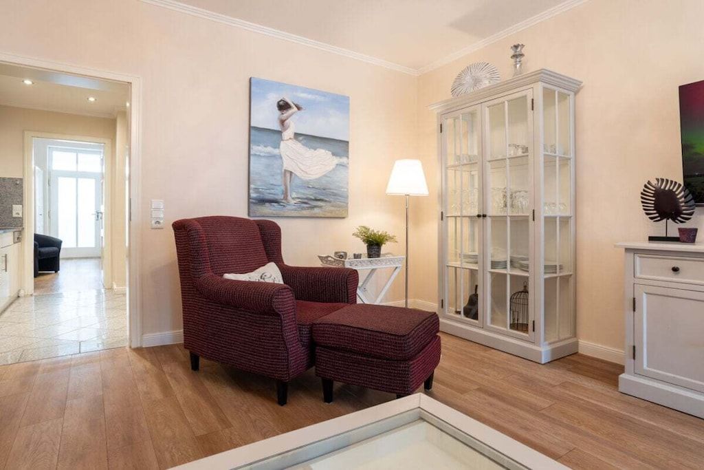 Ein Wohnzimmer mit einem roten Sessel, einer Glasschrank und einem Gemälde einer Frau am Meer.