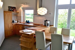 A modern kitchen with wooden cabinets, a dining table with chairs, and a large window providing a view of greenery. - Holiday home in Kosel with swimming pool (Kosel)