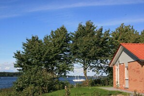 A house with a red roof, a grassy yard, and a lake with sailboats in the background. - Holiday home in Kosel with swimming pool (Kosel)