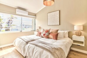 A bedroom with a large bed, a window with a view, a bedside table with a lamp, and a wall-mounted air conditioner. - Stunning 2-Bed Duplex with Air-Con, Close to Beach, Cafés & Restaurants (Cape Town)