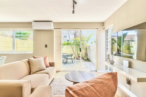 A modern living room with a sofa, armchair, and coffee table. There is a sliding glass door leading to an outdoor patio with a table and chairs. - Stunning 2-Bed Duplex with Air-Con, Close to Beach, Cafés & Restaurants (Cape Town)