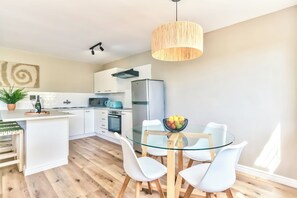 A modern kitchen with white cabinetry, a glass dining table, and a hanging light fixture. - Stunning 2-Bed Duplex with Air-Con, Close to Beach, Cafés & Restaurants (Cape Town)