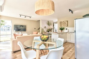 A modern living room with a glass dining table, white chairs, a beige sofa, a flat-screen TV, and a kitchen area with a refrigerator. - Stunning 2-Bed Duplex with Air-Con, Close to Beach, Cafés & Restaurants (Cape Town)