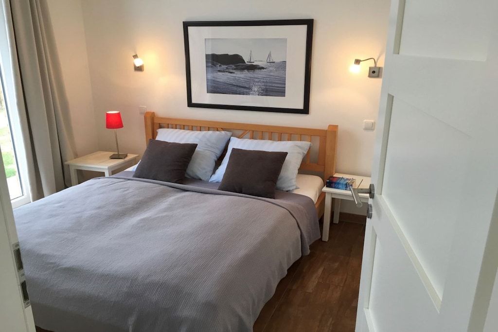 A neatly made bed with a grey bedspread and two pillows, a framed picture above the bed, a red lamp on a bedside table, and a wooden nightstand.