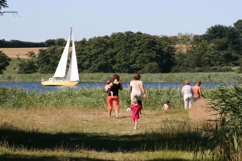 Un groupe de personnes se promène le long d’un sentier herbeux, au bord d’une étendue d’eau, et on aperçoit un voilier au loin.