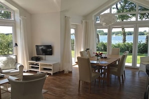 Ein großzügiger Wohnbereich mit einem großen Fenster mit Blick auf ein Gewässer, einem Flachbildfernseher, einem Esstisch mit Stühlen und einer Sitzecke mit einer Couch und Sesseln.