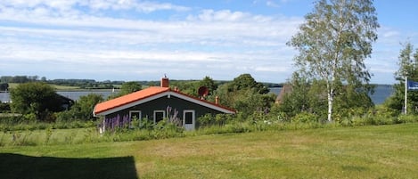 A house with a red roof, surrounded by a grassy field and trees, with a body of water in the background.