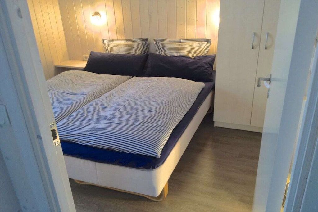 Uma cama de casal com roupa de cama listrada, uma mesa de cabeceira com abajur e paredes de madeira.
