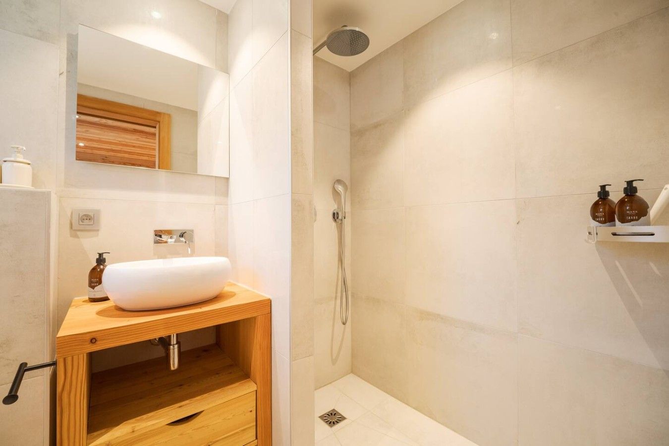 A modern bathroom with a wooden vanity, a white sink, a handheld showerhead, and a shelf with toiletries.