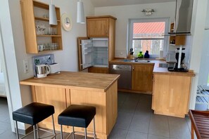 A modern kitchen with wooden cabinets, a built-in island with stools, a refrigerator, and a window with a red-tiled roof view. - Holiday home in Kosel with swimming pool (Kosel)