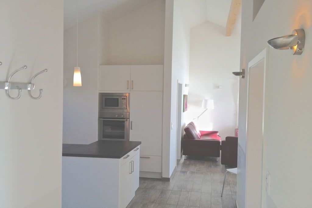 A compact kitchen with white cabinets, a built-in oven, and a microwave. There is a dining area with a red chair and a table.