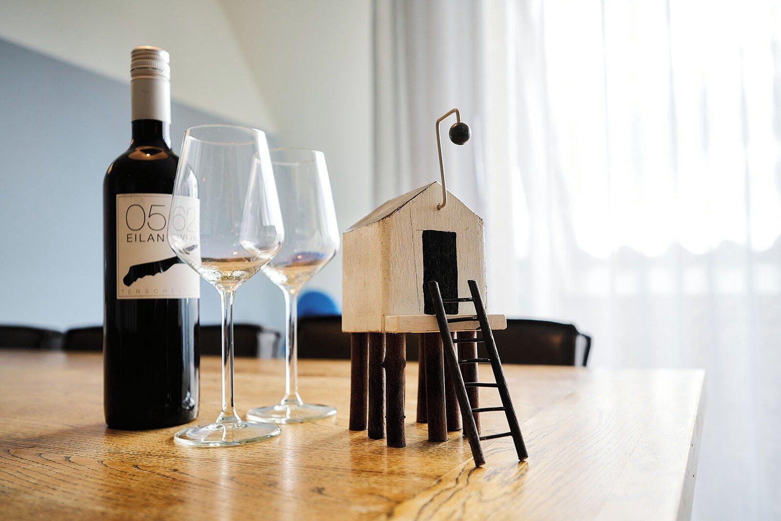 A bottle of wine, two glasses, and a small wooden house model on a wooden table.
