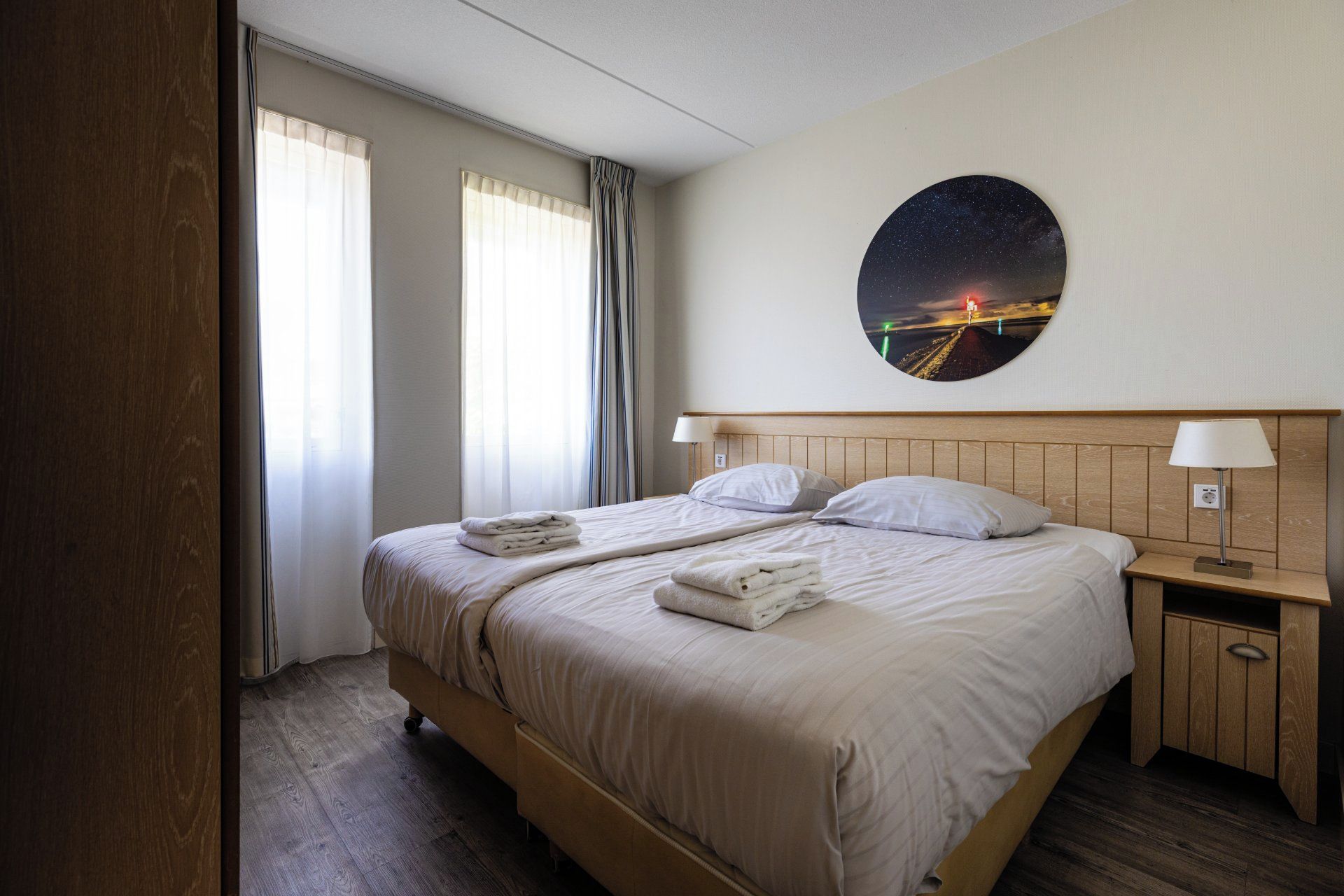 Una habitación de hotel con una cama, dos mesitas de noche, una lámpara de noche, un cuadro redondo colgado en la pared y una ventana con cortinas.