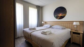 Una habitación de hotel con una cama, dos mesitas de noche, una lámpara de noche, un cuadro redondo colgado en la pared y una ventana con cortinas.