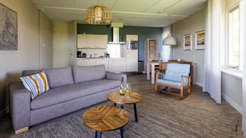 A modern living room with a sofa, two wooden side tables, a dining area with a chair, and a kitchen in the background.