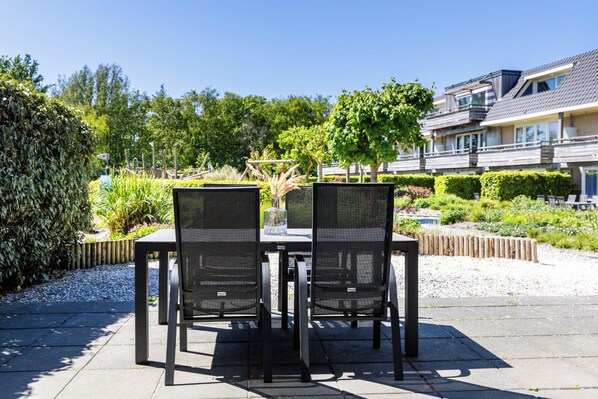 Un área de estar al aire libre con dos sillas y una mesa, rodeada por un jardín con diversas plantas y un edificio al fondo.
