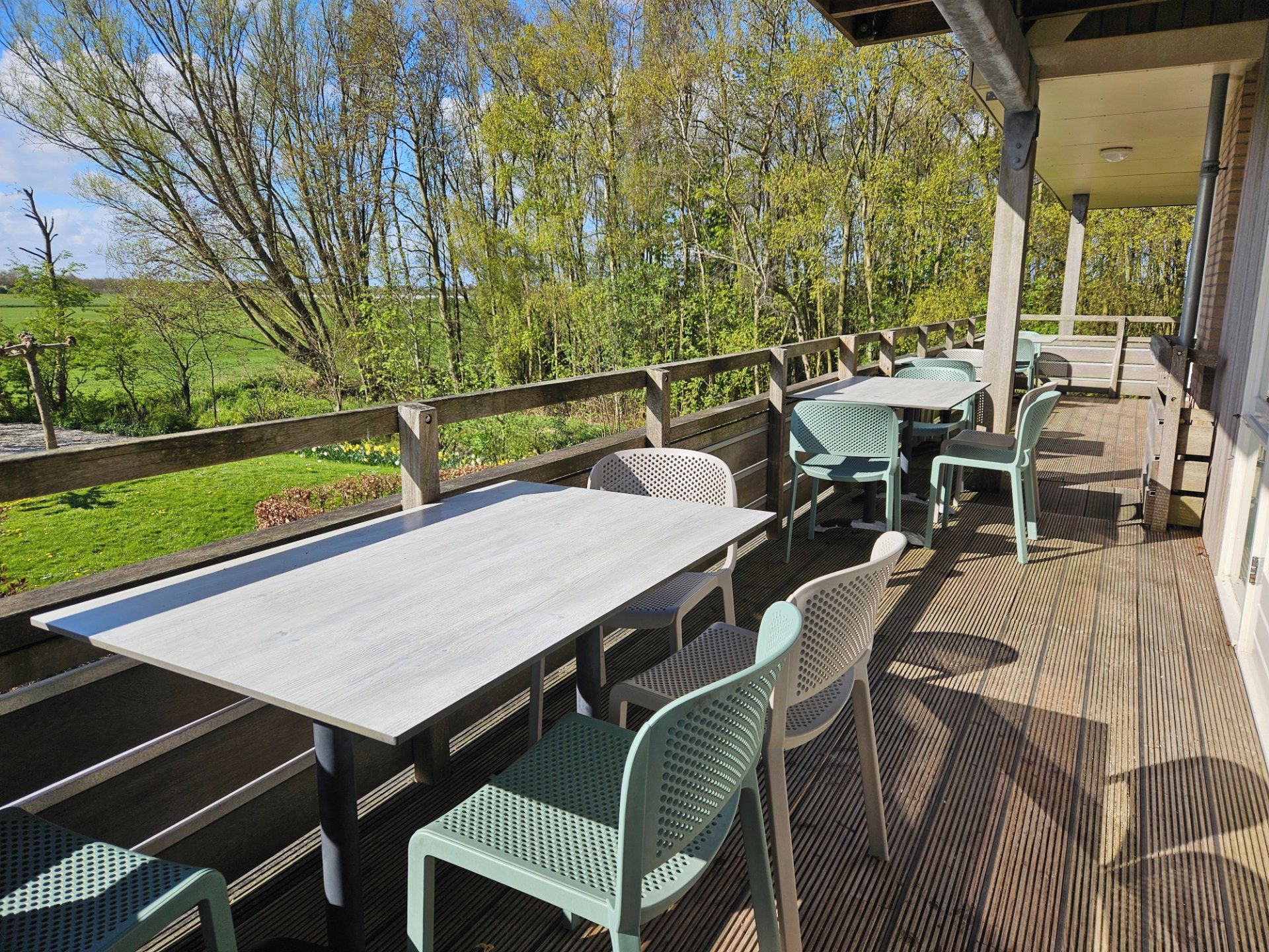 Un patio al aire libre con mesas y sillas, una terraza de madera y vistas a árboles y un campo.