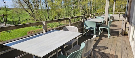 Een terras buiten met tafels en stoelen, een houten terras en uitzicht op bomen en een veld.