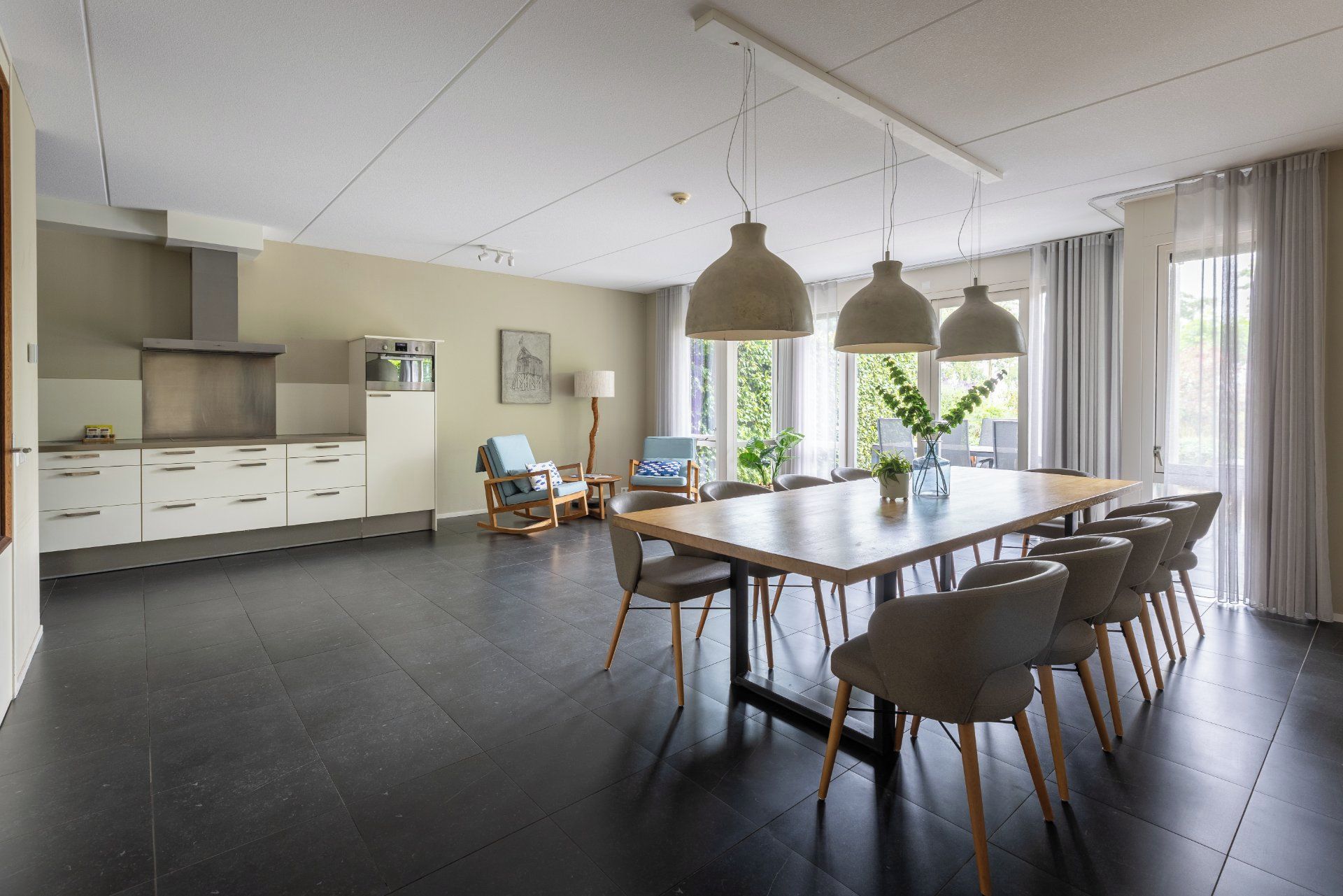 Een moderne keuken met witte kasten, een eethoek met een grote tafel en stoelen, en uitzicht op groen door de ramen.