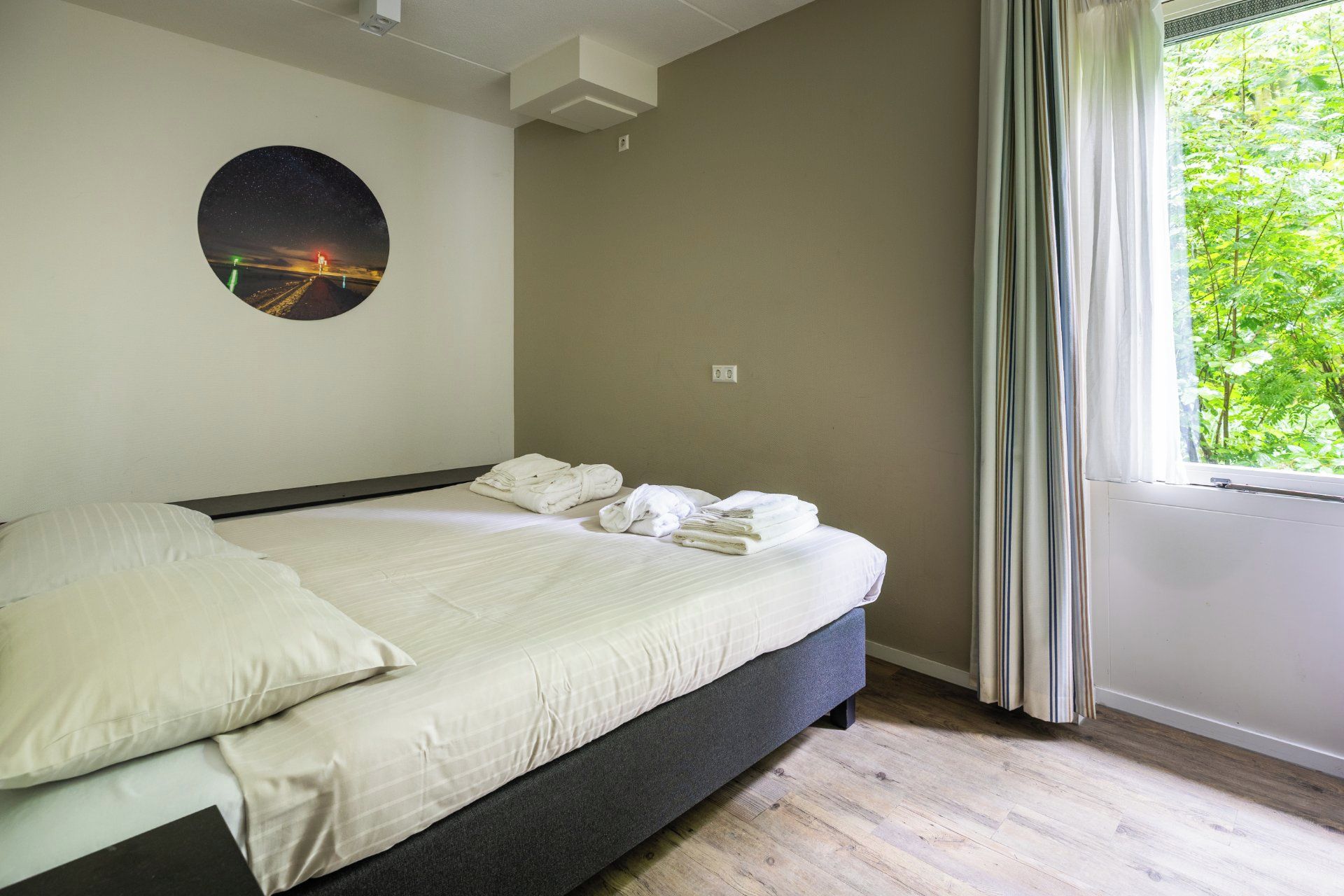 Een slaapkamer met een bed, kussens, een ronde decoratie aan de muur, een raam met gordijnen en uitzicht op groen.