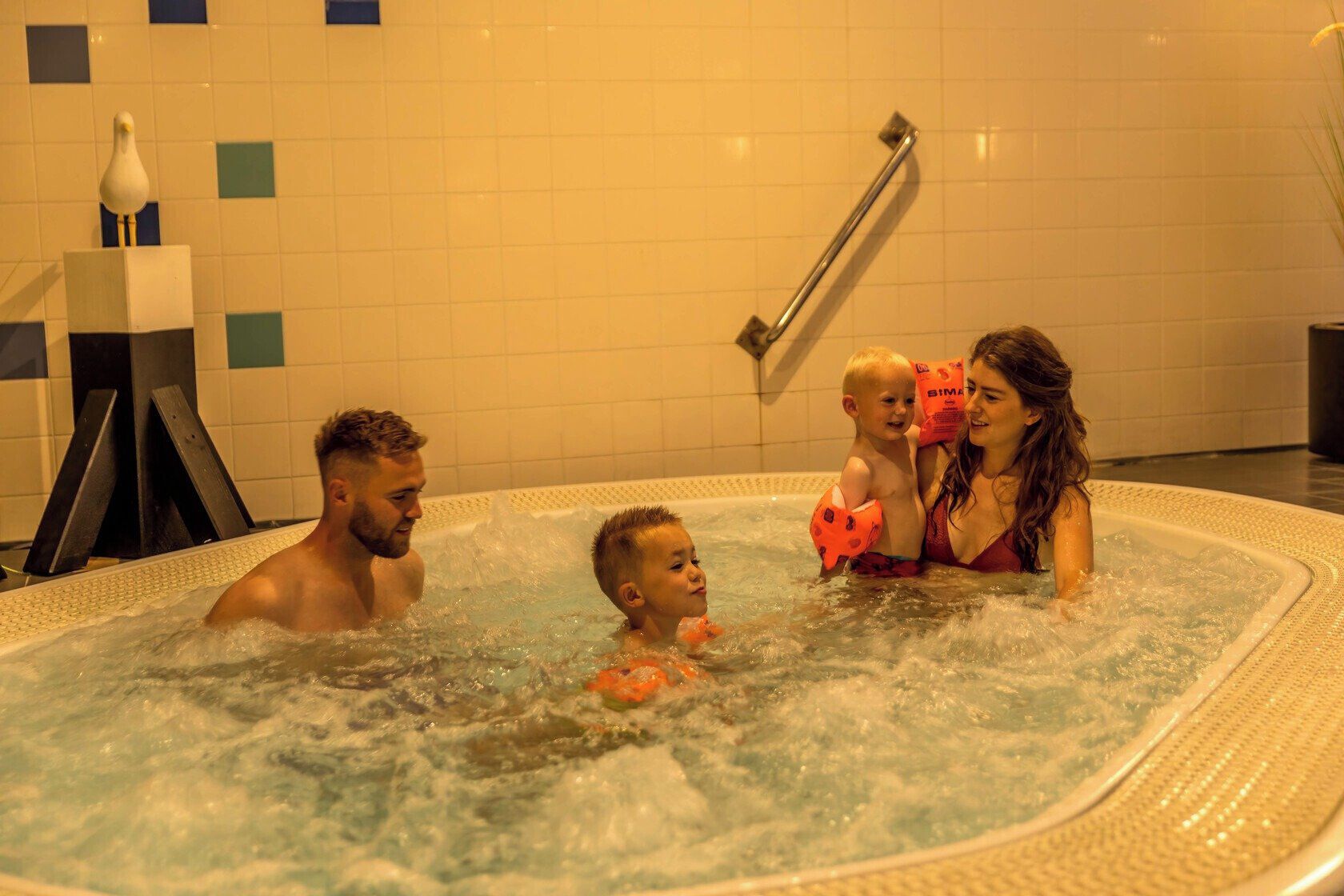 Une famille dans un jacuzzi, avec un mur carrelé et une main courante en arrière-plan.