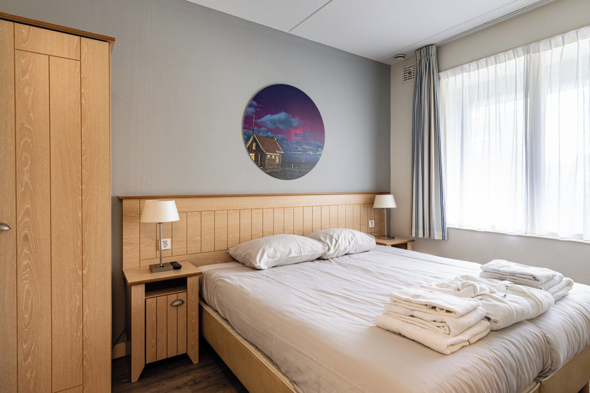 Une chambre d’hôtel comprenant un lit, deux tables de chevet avec des lampes, une armoire et un mur orné d’une œuvre d’art circulaire.