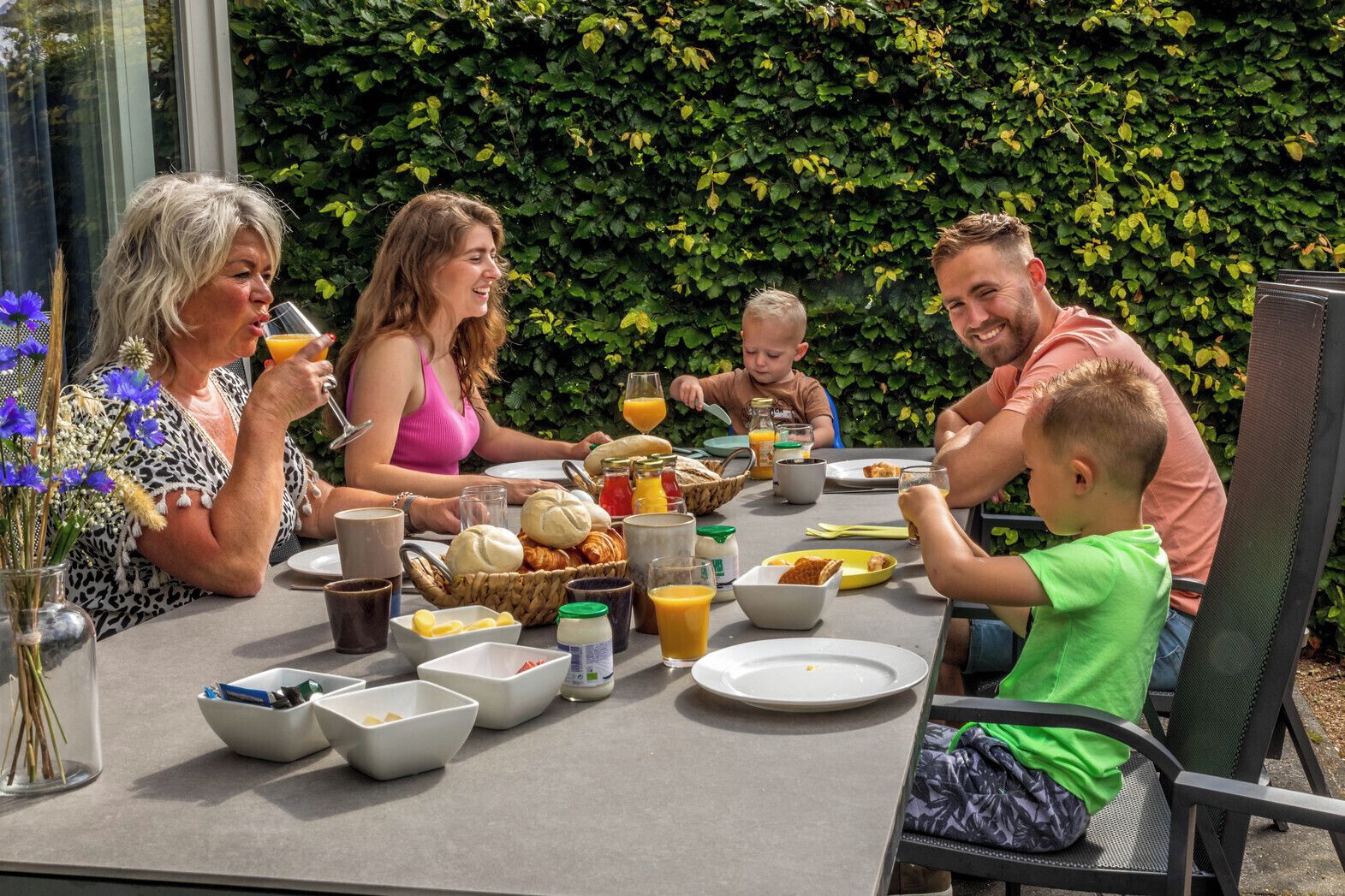 Une famille prend son repas à l’extérieur, autour d’une table dressée pour le petit-déjeuner, dans un cadre verdoyant.