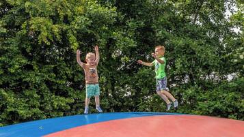 Deux enfants sautent sur un trampoline dont la surface est rouge et bleue, entouré d’arbres verts.