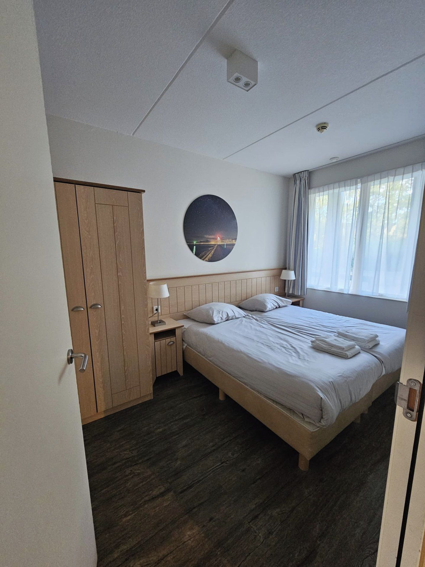 A hotel room with a bed, a nightstand, a wardrobe, a round wall decoration, and a window with curtains.