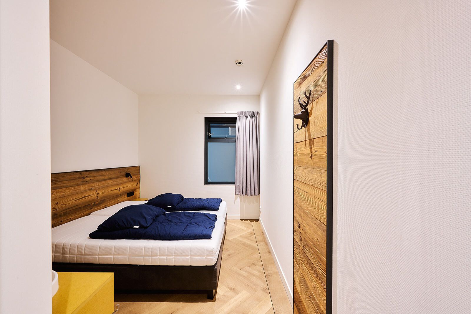 Ein Schlafzimmer mit einem hölzernen Kopfende, einem Bett mit dunkler Bettwäsche, einem Fenster mit Vorhängen und einem Wandbild.
