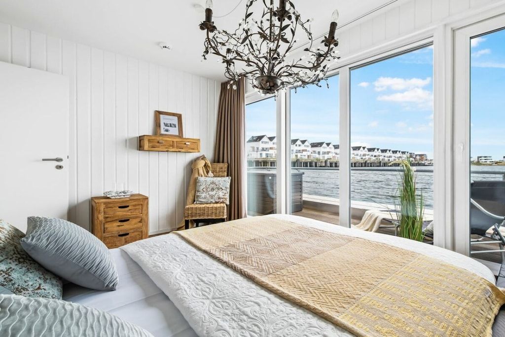 A bedroom with a large bed, a wooden nightstand, a wicker chair, a chandelier, and a view of the water and buildings.