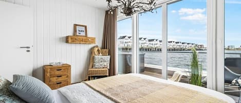 A bedroom with a large bed, a wooden nightstand, a wicker chair, a chandelier, and a view of the water and buildings.