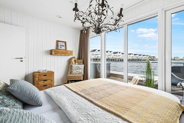 Une chambre avec un grand lit, une table de chevet en bois, un fauteuil en osier, un lustre et une vue sur l’eau et les bâtiments.