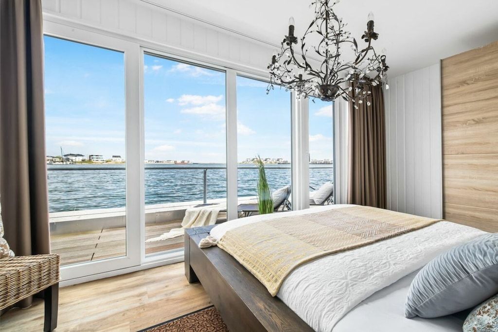 A bedroom with a large window, a bed, a chandelier, and a view of the water.
