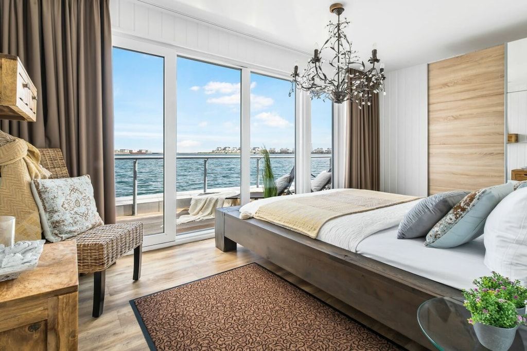 A bedroom with a large window, a bed, a chandelier, and a view of the water.