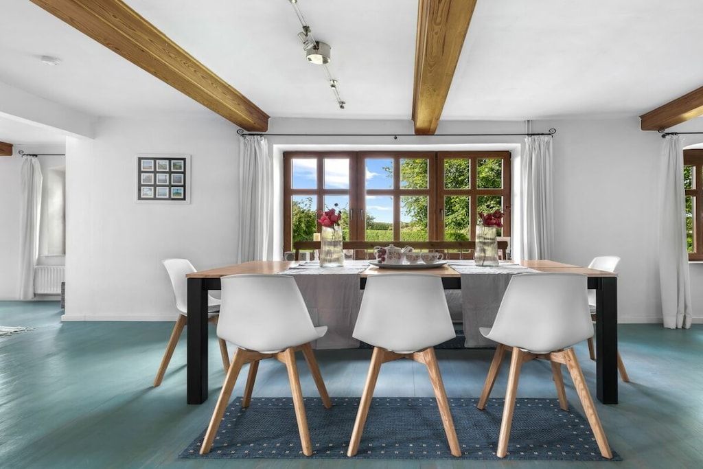A dining area with a table set for four, wooden beams on the ceiling, and a view of greenery outside.