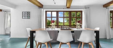 A dining area with a table set for four, wooden beams on the ceiling, and a view of greenery outside.