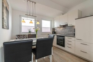 Eine moderne Küche mit weißen Schränken, einem Esstisch mit Stühlen, Pendelleuchten und Blick ins Freie durch ein Fenster.