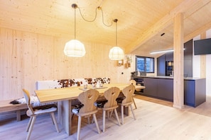 A wooden interior with a dining table, chairs, pendant lights, and a kitchen area.