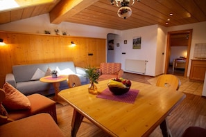 A cozy room with a wooden ceiling, a sofa, a dining table with a fruit bowl, and a small side table. - Holiday apartment with 2 bedrooms (Ruhpolding)